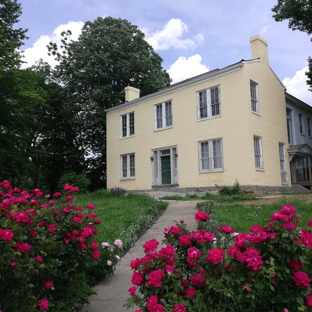 Photograph of a two story brick house painted yellow, with windows that have a white trim, and an evergreen painted front door. In the foreground of the photo are pink flowers.