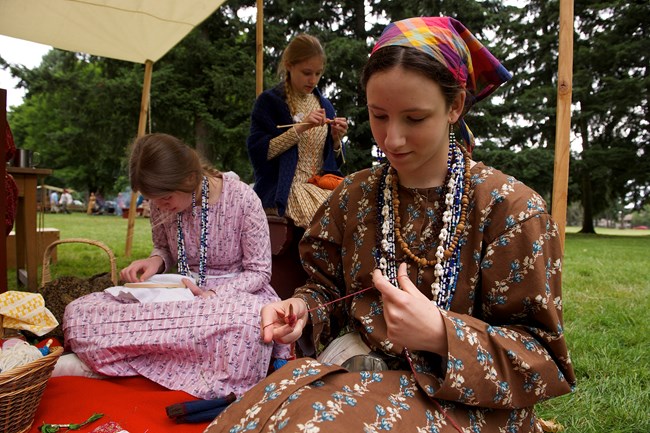 Three girls sit on a blanket working on needlework projects.
