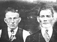 Photographic portrait of two young men with shirt swept-back hair and suits with ties.