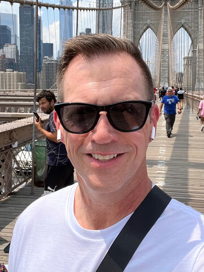 Headshot of Brent Nagen on the Brooklyn Bridge A closeup of a man in sunglasses with people walking and bridge infrastructure in the background.
