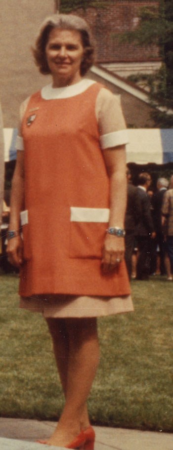 A woman wears a tan dress with a white collar. Over it she wears an orange smock with an NPS arrowhead on the right side.