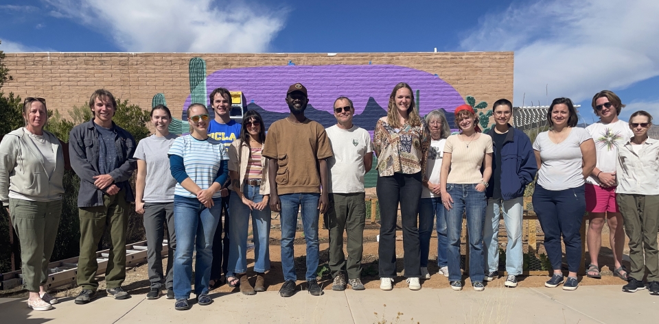 A group photo of a large group of Sonoran Desert Network workers, all lined up around Reagan, an international volunteer. They stand in front of a mural.
