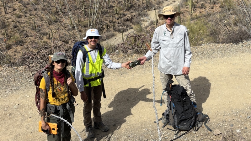 Three people with backpacks standing on a dirt path in the desert, smiling for a photo. Two of the people are holding either side of a wildlife camera.
