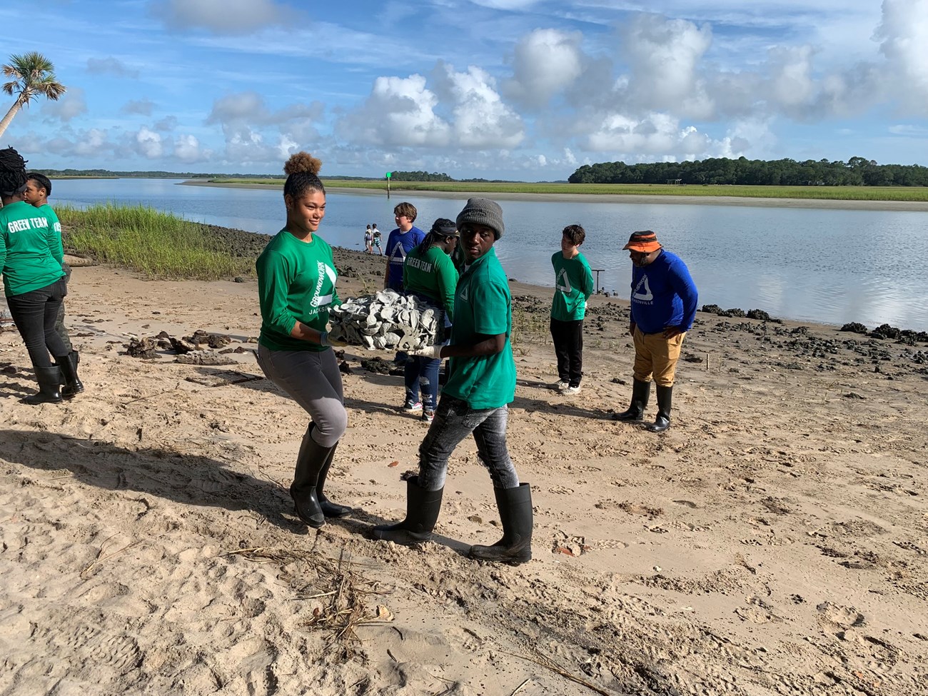 Two people carry block of oysters on shoreline.