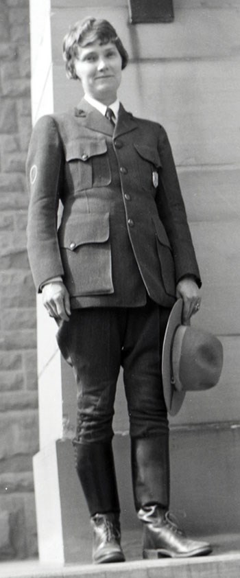 A woman wearing breeches and knee boots stands holding her stetson hat in her left hand.