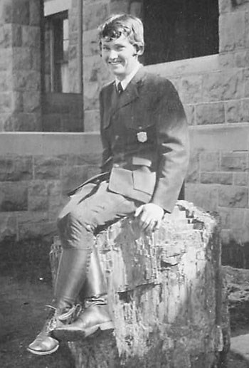 Herma Albertson in the standard NPS uniform with badge poses on a stump of petrified wood in front of a stone building.