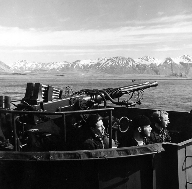 3 Navy crewmembers stand in front of a large double barreled gun platform on USS Casco.