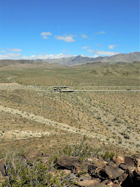 A sparse desert landscape with several hills in the distance. A four lane highway crosses in front of them with a small over pass across a dry gully.