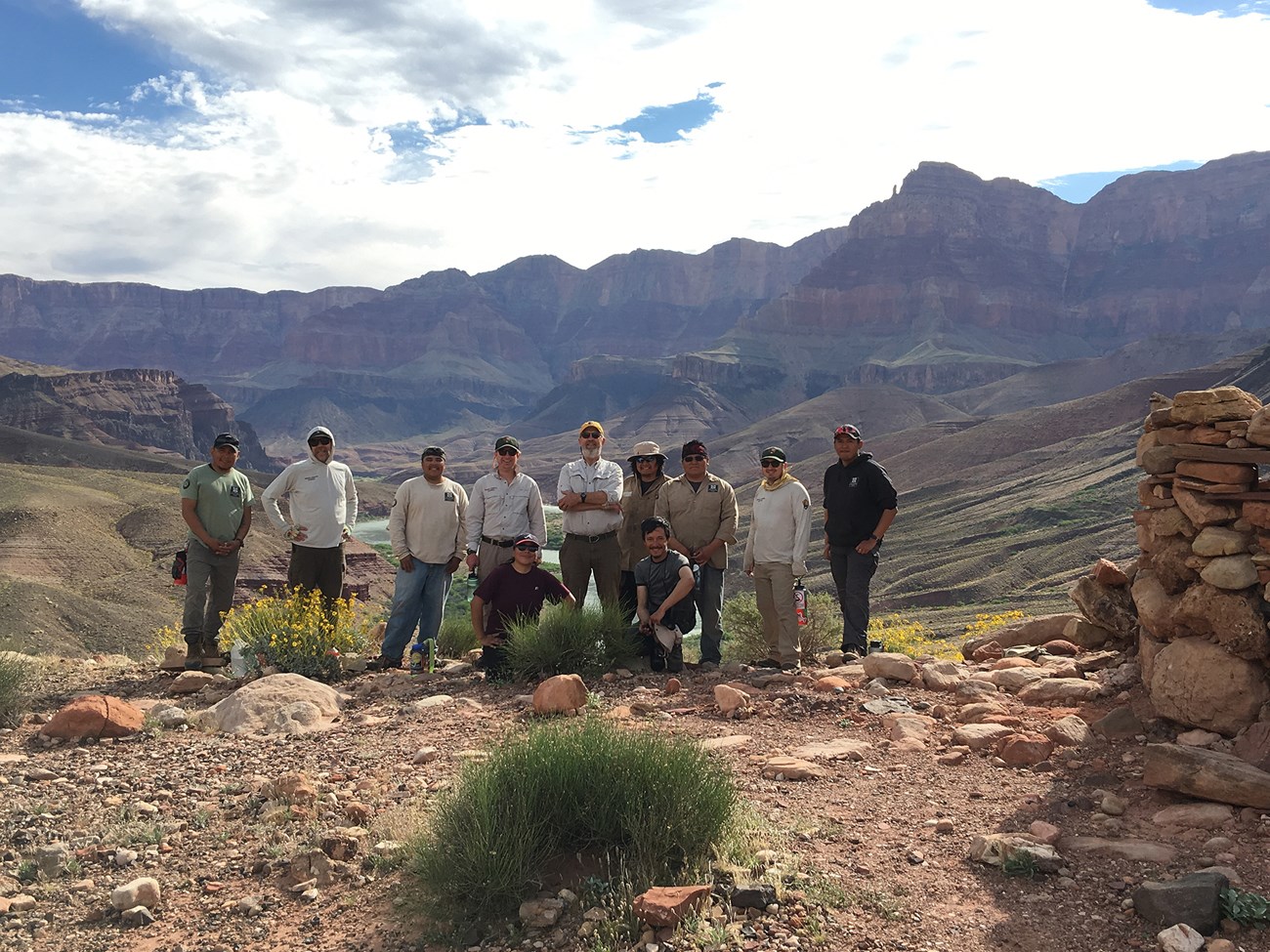 A group of people stand next to a stone structure in front of a landscape of rocks, shrubs, and cliffs.