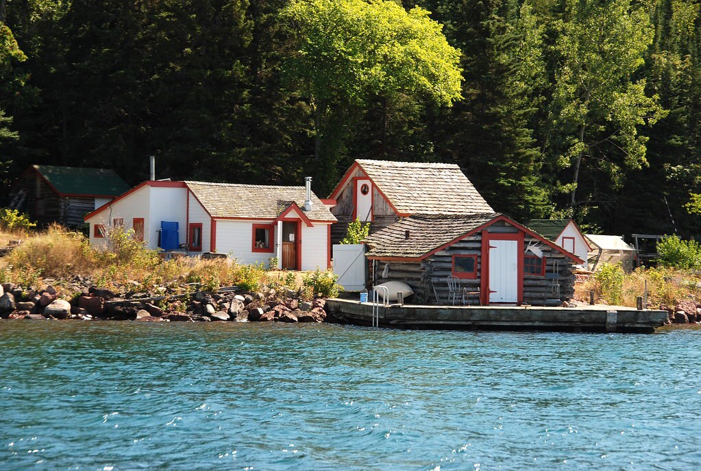 A group of low wood buildings with white and red paint, a dock and a boat sit on the shore of a lake with tall green trees in the background