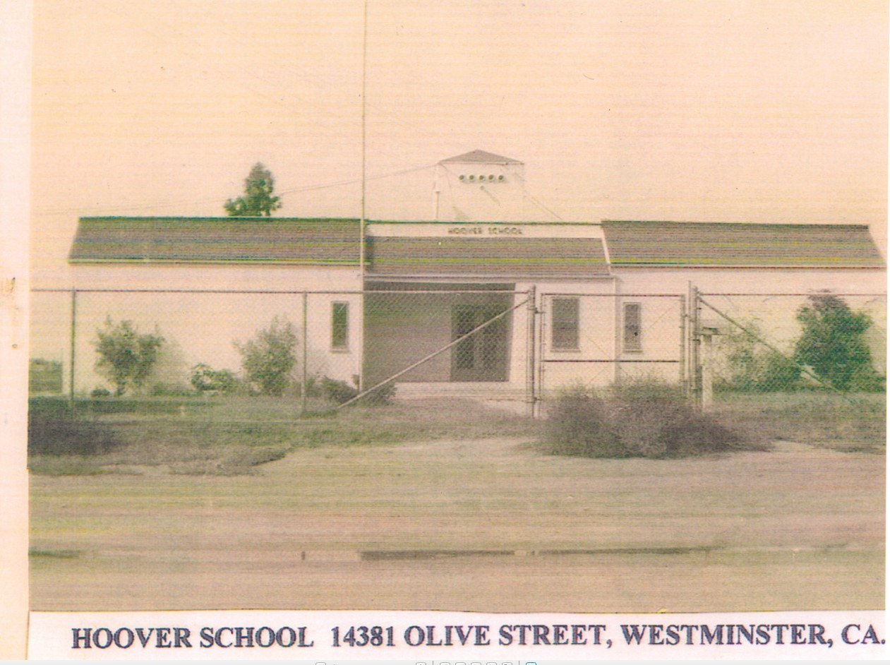 Sepia photo of a one-story white building with three small windows surrounded by chain linked fence.