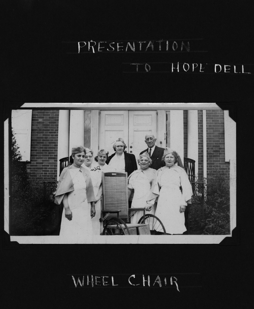 Black and white photo of women in nurse uniform and a man in a suit around a wheelchair