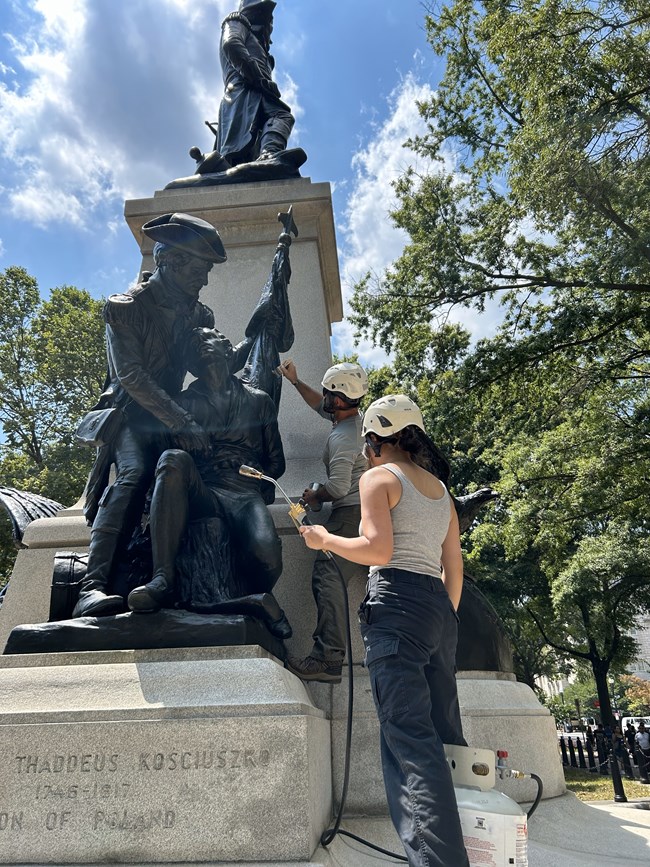 Jessica hot waxing bronze statue Woman holding hot waxing tool and man in background. They are working on the bronze statue of Brigadier General Thaddeus Kościuszko.