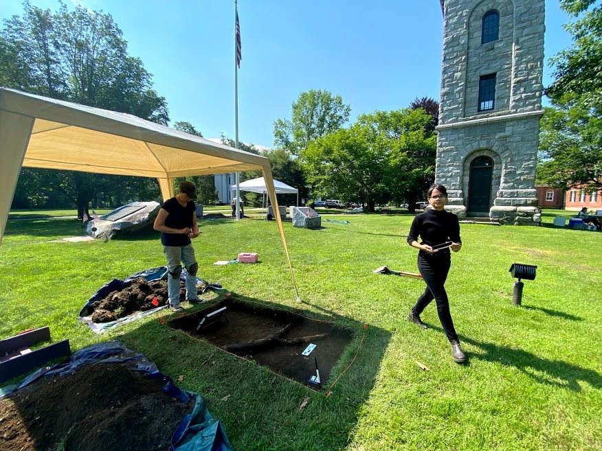 Stockbridge-Munsee Community Band of Mohicans Archeology Project in Upper Housatonic NHA Small, rectangular dugout archaeological tract within large town green. Person stands to left of tract with archaeological materials under tent; another person passes by to the right. Large stone tower and trees in background.