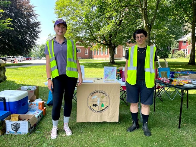 Two people stand smiling in neon safety vests outside on town green, in front of table with various materials, maps, and snacks. A small banner on the table reads "Mohican Nation Historic Preservation" around tribal insignia.