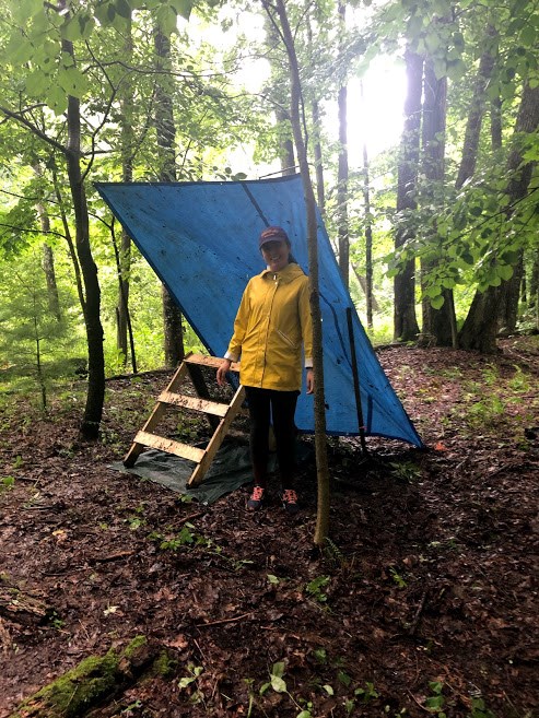 Woman stands in yellow raincoat and boots under angled tarp and project materials in wooded area