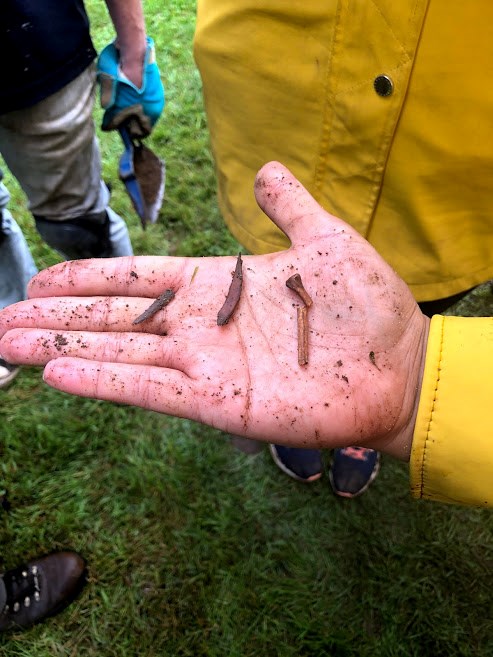 Closeup of hand, palm open and face up, with three small, rusted metal objects, including one broken metal nail. Another hand carrying small hand shovel in background.