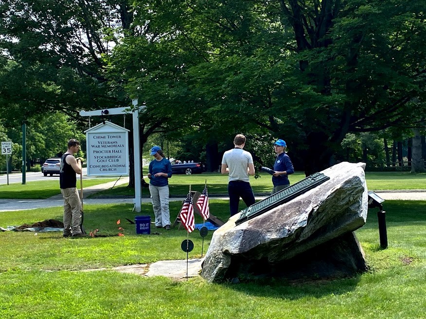 Four people stand on town green amidst archeological project materials. Large rock with plaque in foreground, and white historic town sign in back.