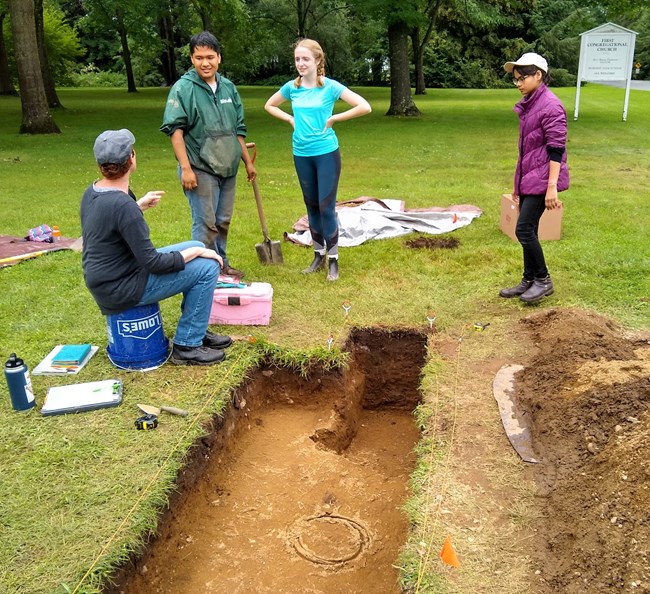 Three young people stand, one with shovel in hand, in front of small dug-out archaeological tract revealing a rectangle of dirt within green grass. They appear attentive, listening to seated man with various archeological tools. Large dirt pile at right.