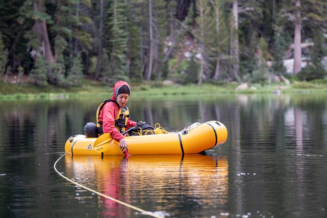 Field scientists wears hooded red jacket and life vest and collects water quality data from a yellow raft in the middle of a mountain lake.