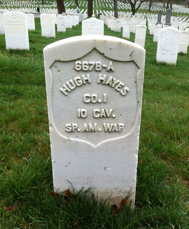 White marble slab with text on it saying "6678-A - Hugh Hayes - Co. 1 10 Cav. Sp. Am. War." Many similar marble stones are behind the gravestone