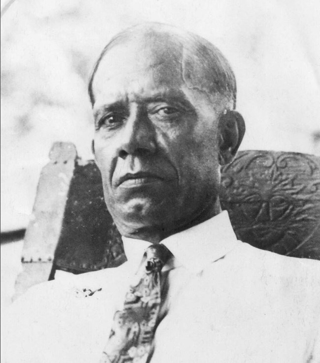 Black and white photo of African American man wearing a white shirt and spotted tie sits in a wooden chair.