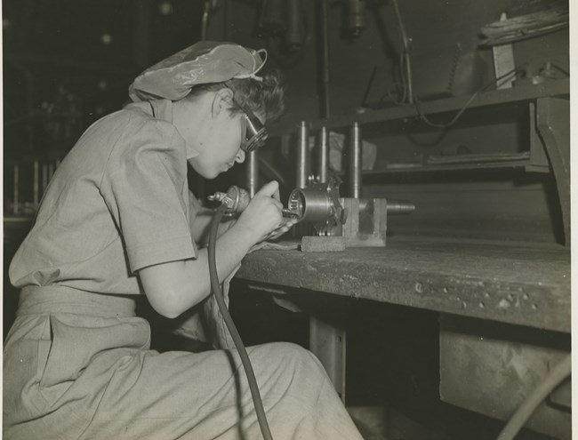 Black and white photo of a woman in pants and a hairnet bent over a small metal machine