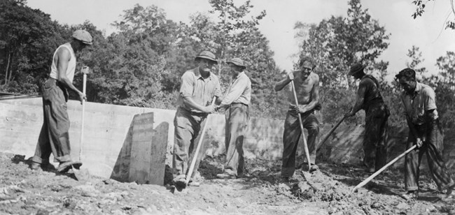 A black-and-white photo of six men working with shovels around an exposed concrete foundation.
