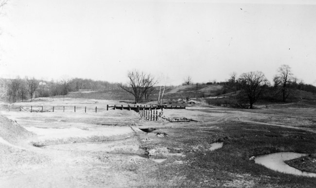 A black-and-white photo of a pier under construction; what will be the lake is still a grassy field.