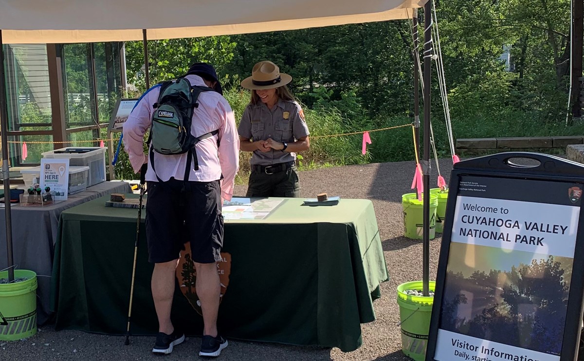Uniformed ranger smiles and talks to a visitor under a tent; they stand on either side of a table with green tablecloth; to the right, a sign that says "Welcome to Cuyahoga Valley".