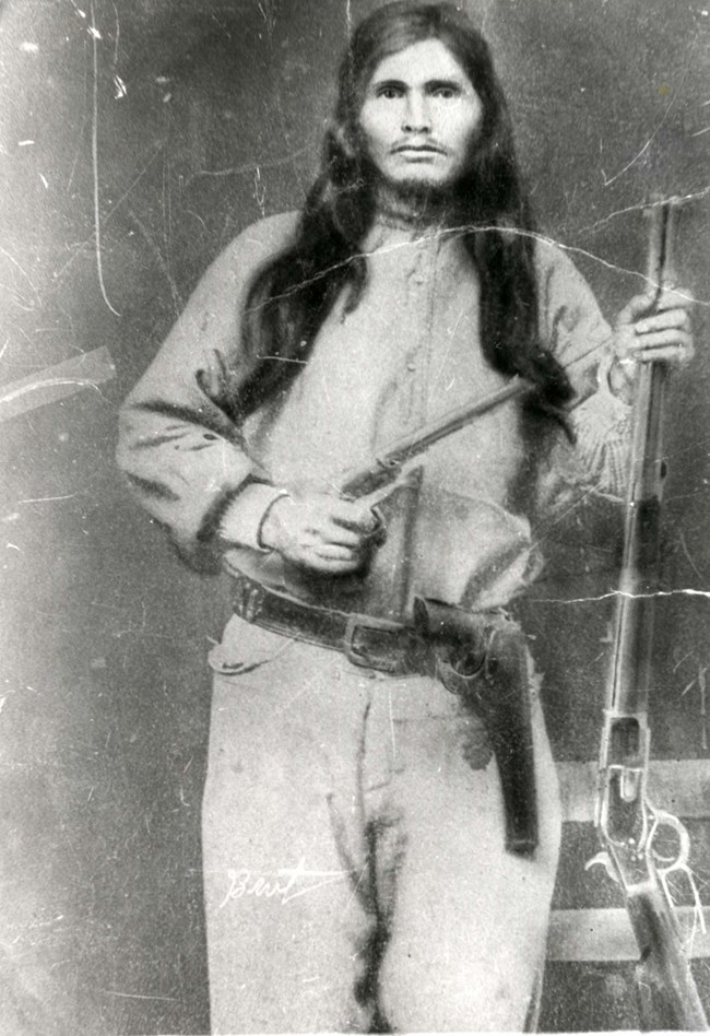 Ned Christie Historic photo of Ned Christie, a man with long dark hair standing holding two guns with a serious expression.