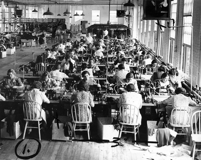Women sewing at long tables next to tall windows in a garment factory.