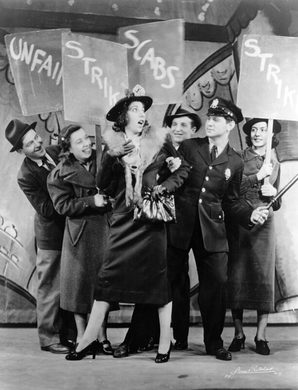 A woman sings on stage surrounded by actors holding signs that read "Strike," "Scabs," and "Picket."