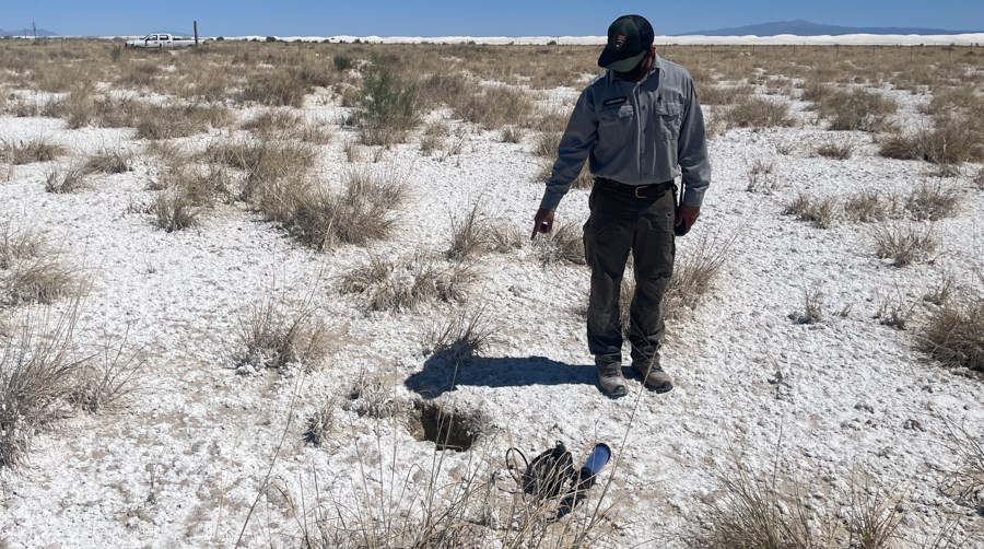 EC-30 Spring Person pointing at the spring orifice in a small depression in the ground in a flat landscape covered in white sand and many scattered, dried grasses.