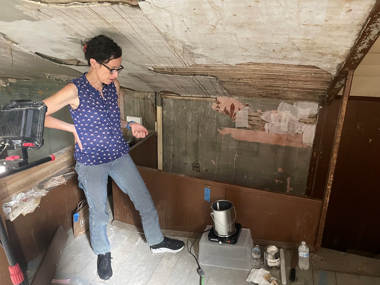 An NPS conservator prepares materials to remove historic plaster and stenciling, visibly crumbling on the wall behind her.