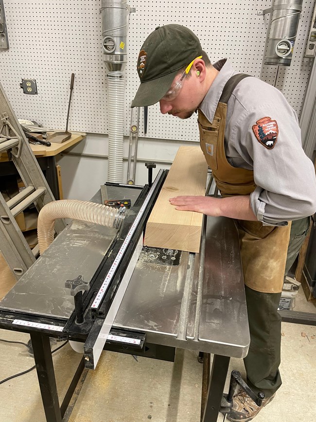 A person wearing an NPS uniform, ear, and eye protection uses a table saw to cut a wooden plank.