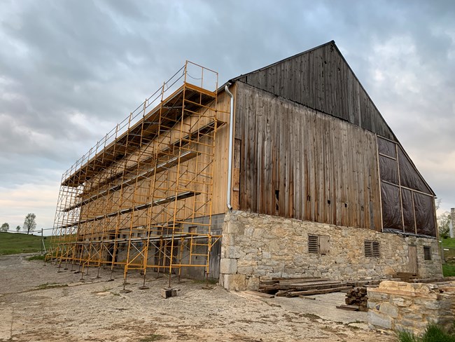 Construction scaffolding covers the side of a large historic barn. The barn is timber framed with a stone basement.