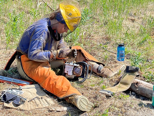 A woman in wildland firefighter gear sits on the ground working on a chainsaw