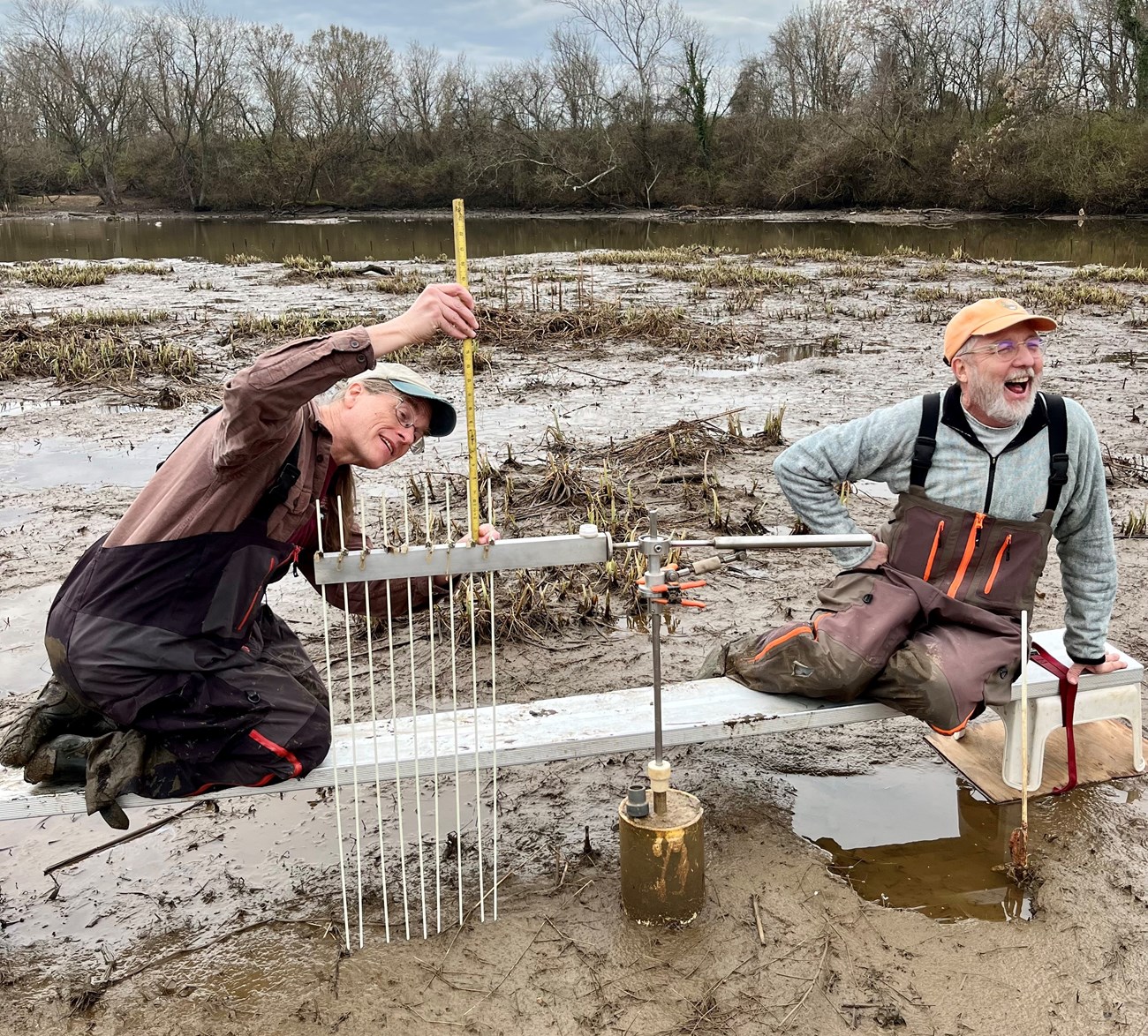 Two ecologists sit on a bench in the swamp, one measuring with SET, the other laughing toward the camera.