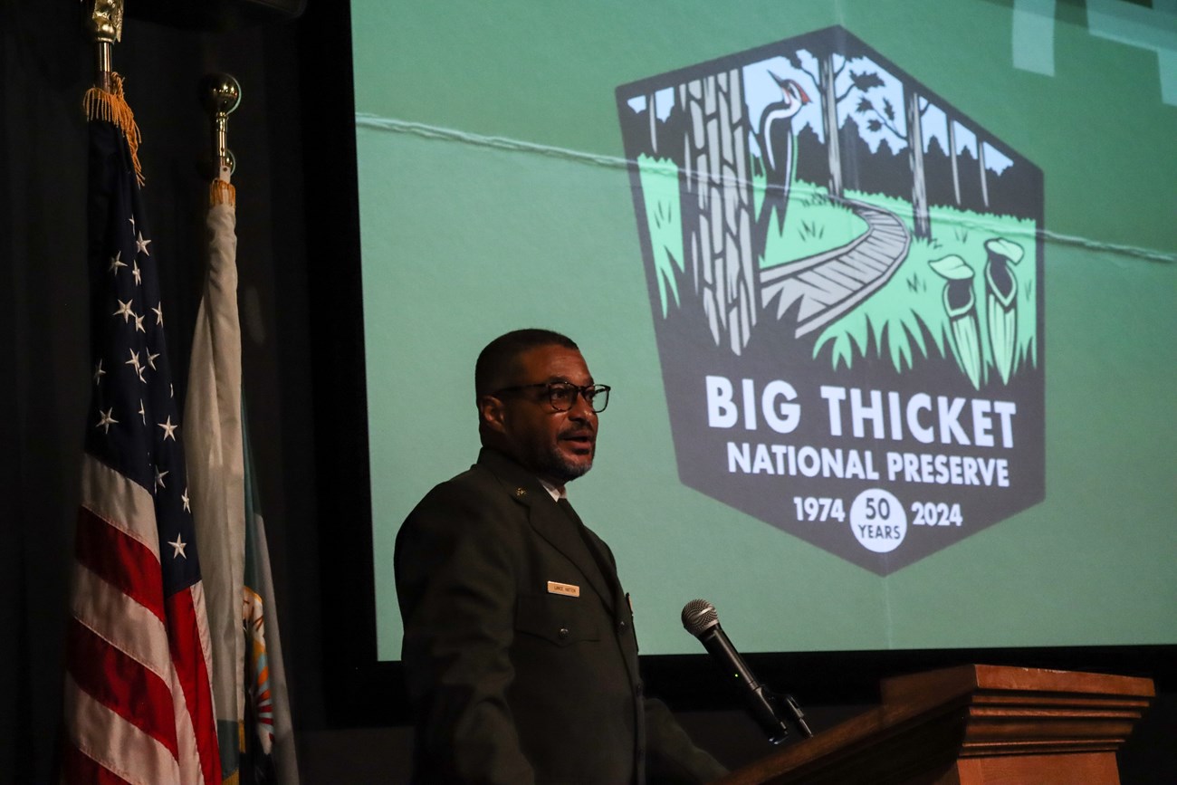 DRD Hatten a park ranger speaking at a podium in front of a screen that says Big Thicket National Preserve 50th anniversary.
