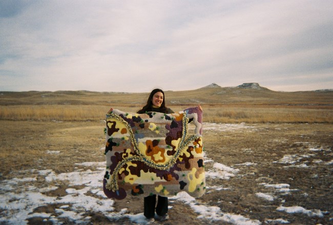 A woman holds up a colorful rug in the middle of a desert.