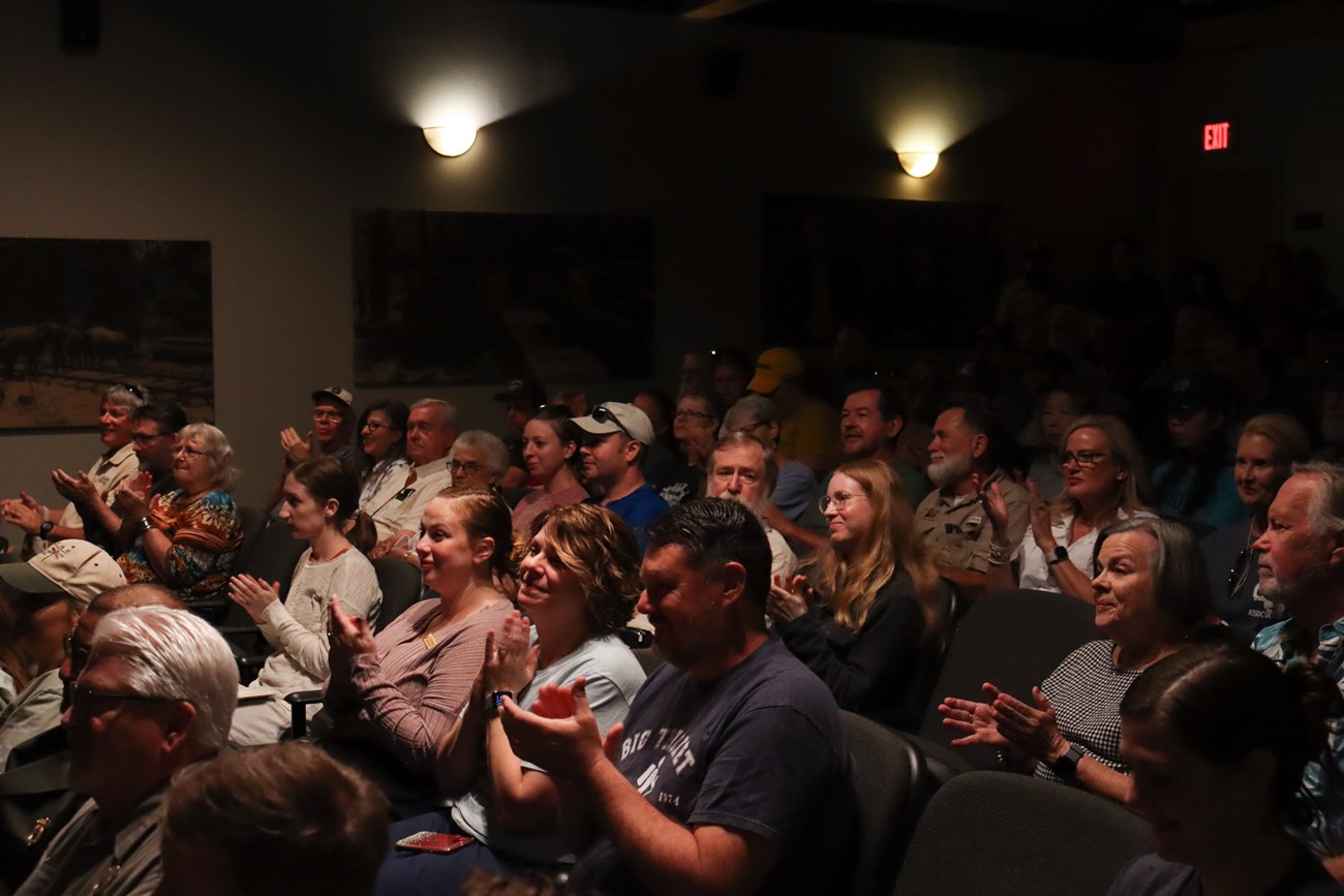 A seated audience in a theater clapping their hands following a speech.