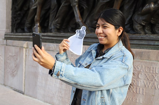 Young woman stands with a cut out of Harriet Tubman in front of a Civil War monument and takes a selfie.