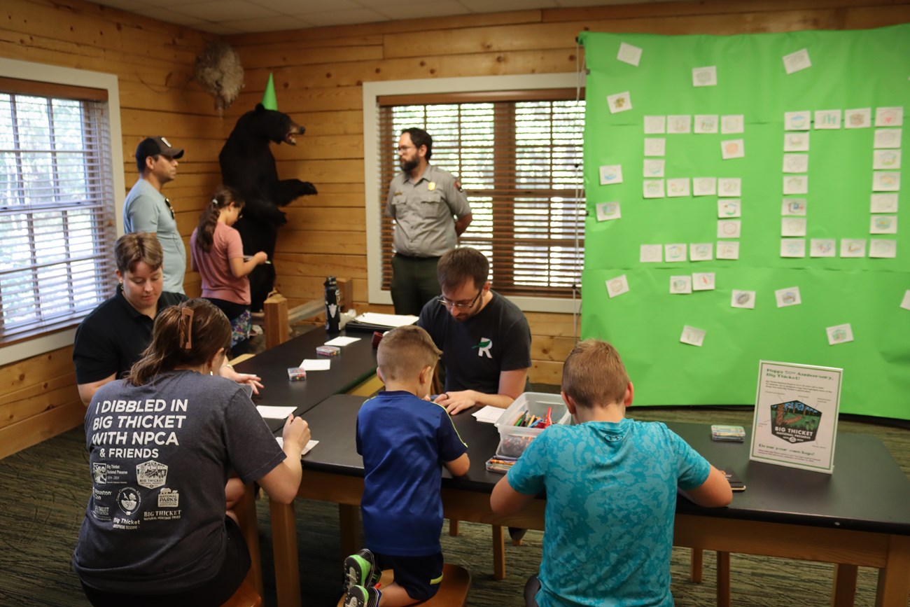 visitor center activities people seated at an activity table inside a room with wooden walls, with a park ranger standing next to a taxidermy bear.