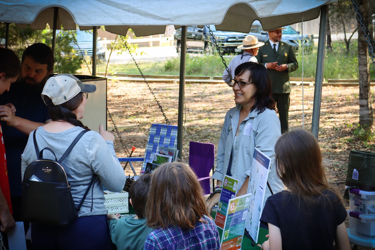 NPCA table a woman at a table under a tent talking to a family with small children. Flyers and maps are set up in front of her. A parking lot is in the background.