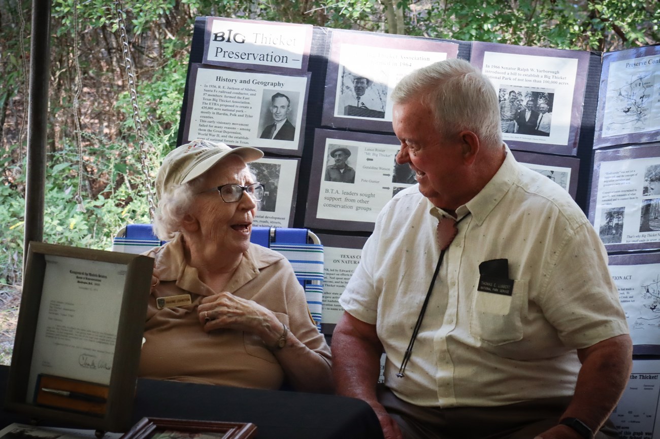 Maxine and Thomas an older woman sitting next to an older man at table while sharing a laugh. Behind them is a display board with historic photos and text.