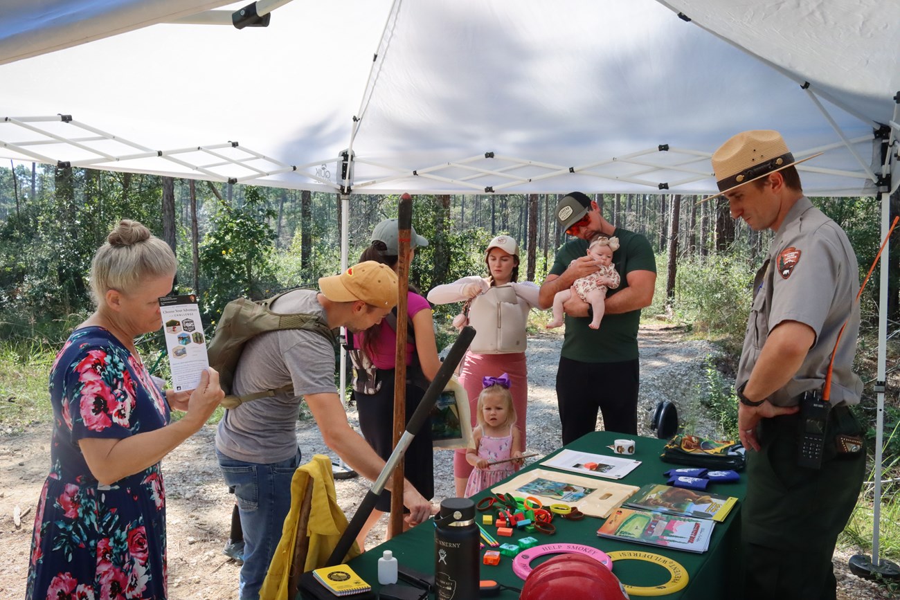 Booth on the Longleaf Pine Trail a park ranger standing at a table full of colorful props and objects, talking to a family with small children and adults.