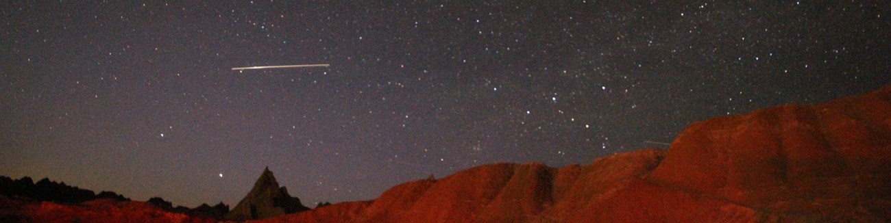 a starry night sky with a white streak in the left corner, appearing just over some red badlands buttes