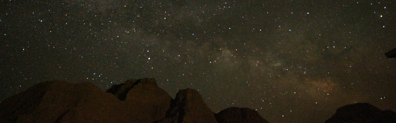 a starry night sky with a cloudy streak appears above badlands buttes