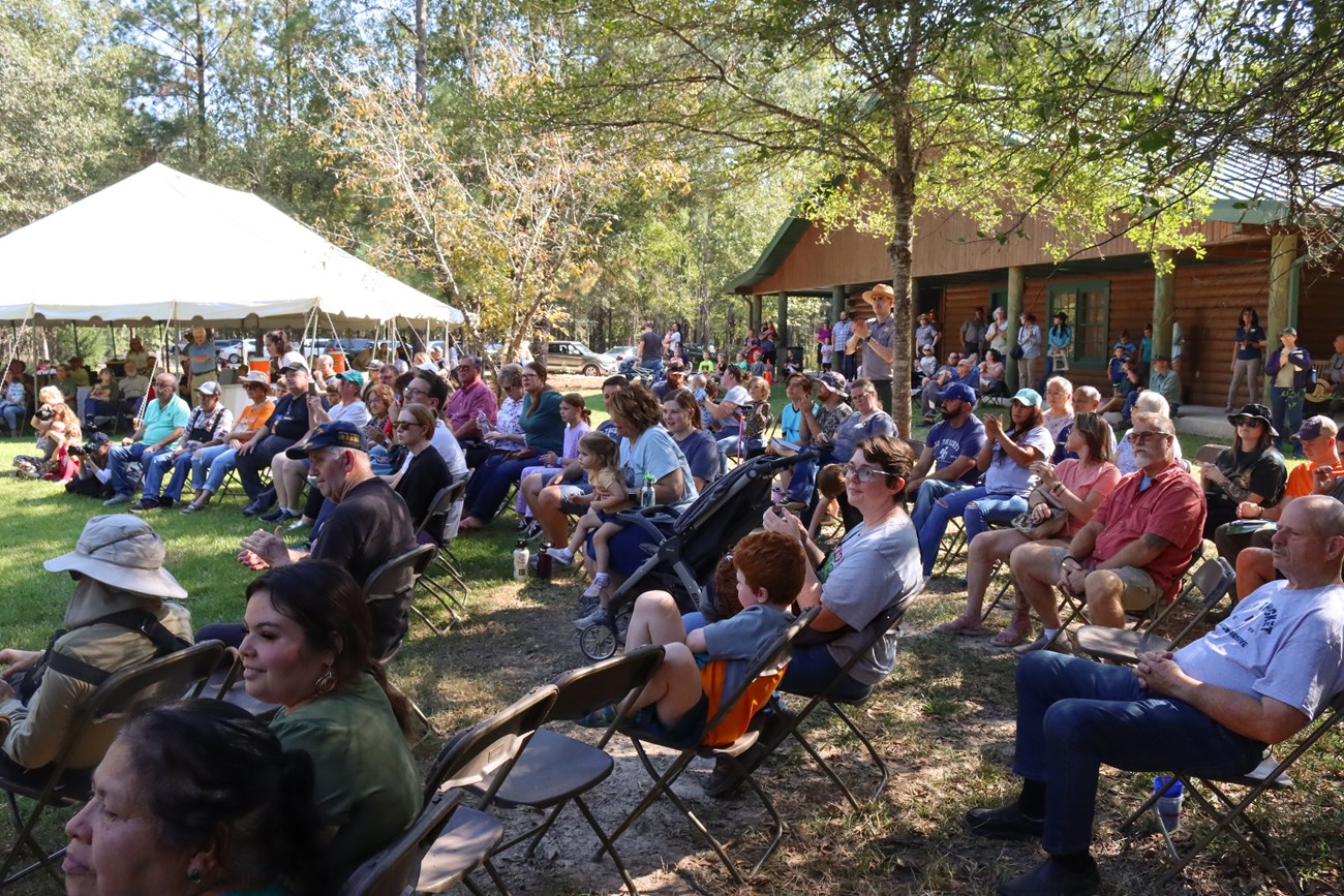 an audience of about one hundred people seated outside on a lawn near a big white tent and visitor center building.